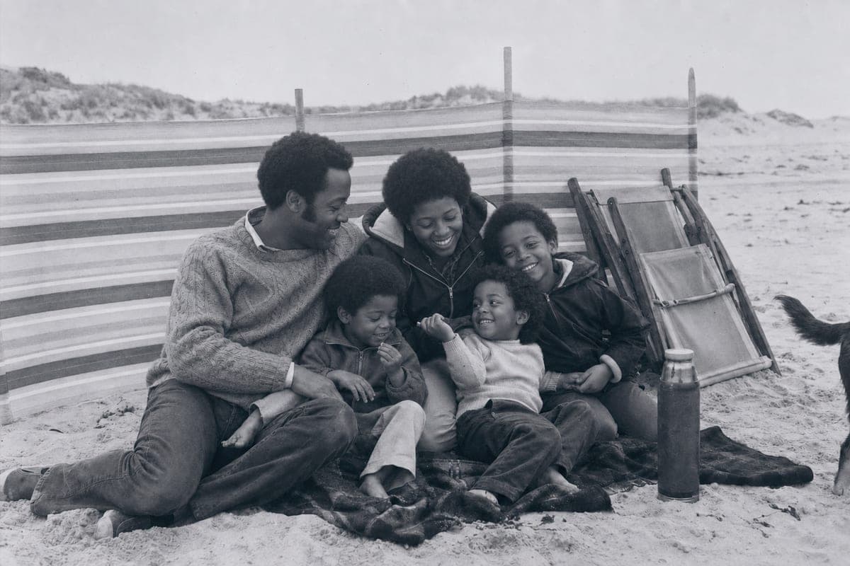 A family of five on a blanket behind a striped windbreak on a pale beach, in black and white