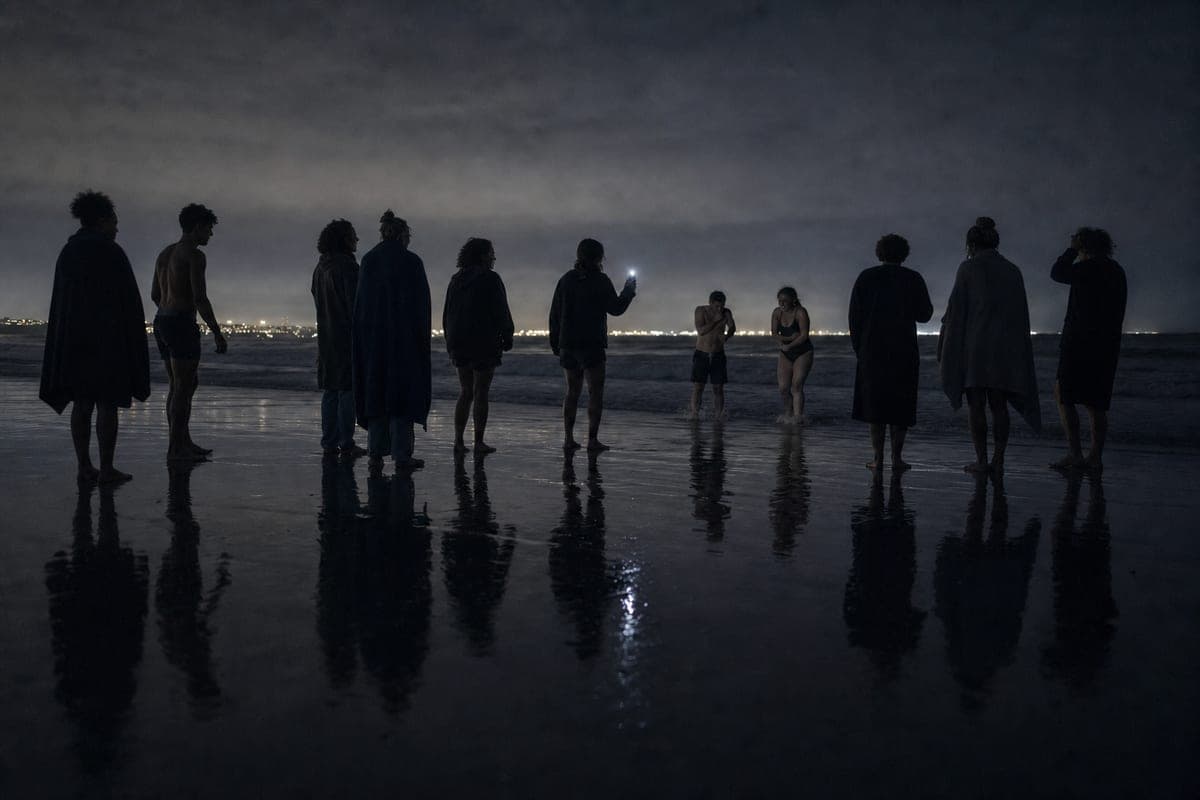 A line of people on wet sand at night, silhouetted against distant town lights, two of them already in the shallows